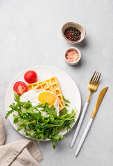 Waffles with fried egg, cherry tomatoes and arugula on a white plate on a gray texture background with spices.