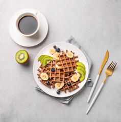 A freshly prepared chocolate waffles with kiwi,  blueberries and banana slices on a white plate with a cup of coffee on a light gray background.