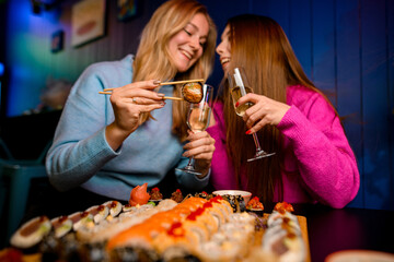 Two cute happy women happily eating sushi rolls with chopsticks and holding glasses of champagne