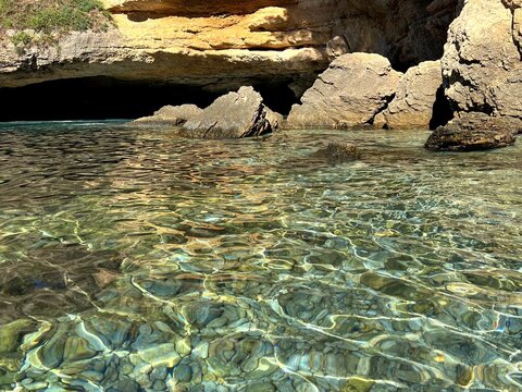 Sea clear water with smooth stones and pebbles by rocky coast with cave. 