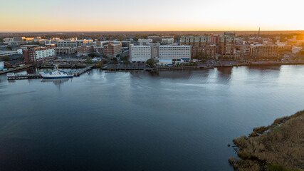 Obraz premium Dawn aerial shot of Wilmington, North Carolina, with Cape Fear River and moored boats illuminated by sunrise, showcasing the peaceful waterfront cityscape.