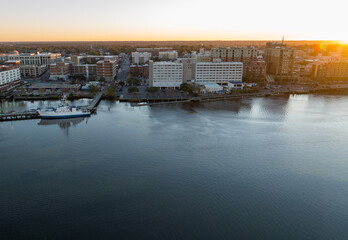 Dawn aerial shot of Wilmington, North Carolina, with Cape Fear River and moored boats illuminated by sunrise, showcasing the peaceful waterfront cityscape.
