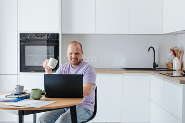 A white man, 35-39 years old, in a lilac T-shirt, works at a laptop in a bright kitchen. He holds a cup of coffee.
There are papers and cups of coffee on the table.