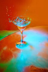 Ice cube falling into glass with martini cocktail against orange background in neon light with...