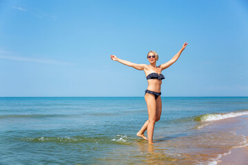 Laughing woman in a black bikini on the seashore. Active lifestyle, travel and relaxation at the resort on a sunny day. Full height.