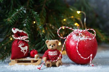 Christmas card with a toy bear, a red apple and a bag of gifts on the table, fir and pine branches with a light garland