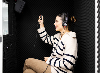 A grown girl is inside a soundproof audiometry booth. Hearing loss treatment. Young woman with positive expression wearing audiometric hearing aids while listening to tests.