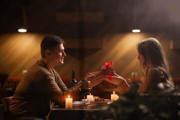 Happy young couple celebrating Valentines day having romantic dinner at home. Loving man giving gift box his woman - obrazy, fototapety, plakaty