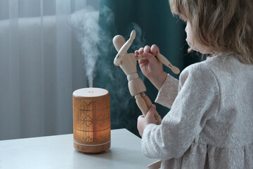 Wooden diffuser, yellow light, electric aromatic. A little blonde girl next to a wooden mannequin. The concept of care and comfort, air humidification in the house. - obrazy, fototapety, plakaty