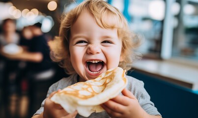 Cute baby with blond hair with a crepe on Mardi Gras, holding a mouth-watering pancake, laughing with delight, little greedy boy or girl with open mouth, smiling, thrilled and eager to taste the snack - obrazy, fototapety, plakaty