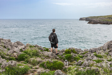 Fototapeta na wymiar Back view of a mature man with backpack and casual clothes looking at the sea with his dog in nature. Travel and nature concept