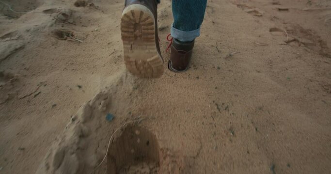 Camera follows woman or man in brown heavy duty leather boots walk on dry sandy surface. 
