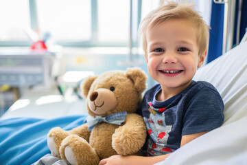 Small young boy smiling in hospital room with brown teddy bear on his lap.