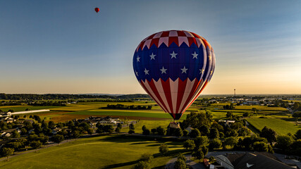 An Aerial View on a Stars and Stripes, Hot Air Balloon Floating Over a Countryside Community, on a...
