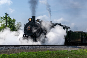 A View of a Narrow Gauge Restored Steam Passenger Train Blowing Smoke and Lots of Steam, Starting To Pull Out of a Station on a Summer Day
