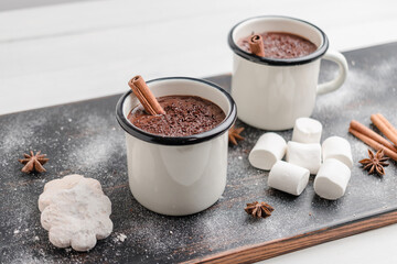 Homemade spicy hot chocolate drink with cinnamon stick, grated chocolate in enamel cup on wooden table with cookies, white marshmallows and star anise