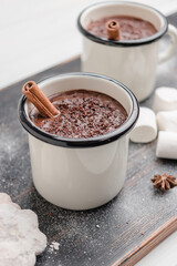 Homemade spicy hot chocolate drink with cinnamon stick, grated chocolate in enamel cup on wooden table with cookies, white marshmallows and star anise