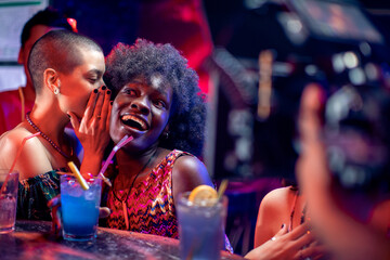 Surprised young woman listening to friend whispering in her ear at bar