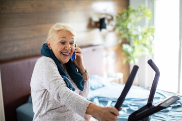 Happy senior woman having phone call on home fitness bike