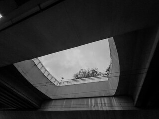 Black and white photo of a concrete underpass, taken from below. The underpass frames the sky and...