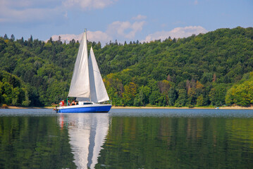 Three people are sailing on a blue sailboat. Sailing a yacht on Lake Solina in summer. Panorama of the Bieszczady Mountains, Poland.