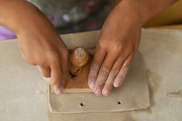 Child's hands in pottery-making process, pressing wooden tool into soft clay, creating piece of...