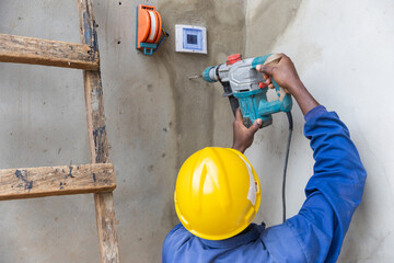 Back photo of the helmeted electrician using the drill to make a hole in the wall