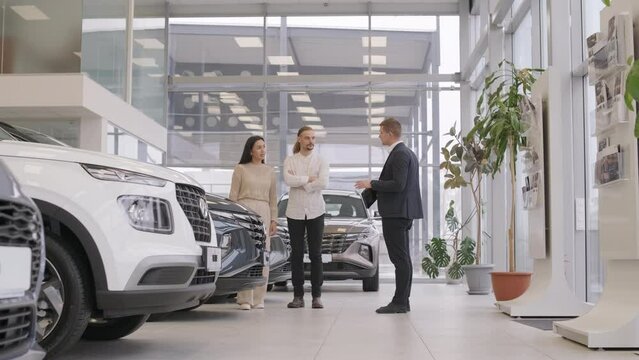 Young car salesman showing to young couple new automobile at dealership salon.