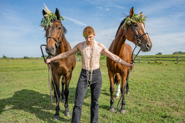 Jeune homme puissant torse nu tenant deux chevaux avec des colliers de fleurs - obrazy, fototapety, plakaty