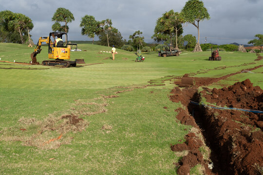 Man and construction equipment digging trenches through green grass of a golf course to install irrigation and utility lines, Poipu, Kauai