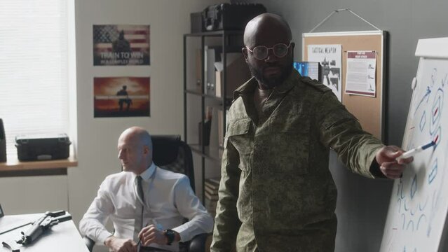 Medium long side shot of African American male cadet standing near whiteboard explaining chosen tactics to Caucasian military professor during class in academy