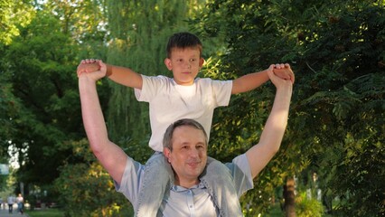 Pleased father has son sitting on shoulders among trees in city park. Grateful father strolls in city park with boy perched on shoulders. Father and satisfied child relish stroll in city park