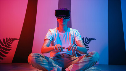Smiling teenage programmer boy sitting on the floor with a virtual reality headset on, making hand gestures, typing, programming and scrolling in the air.