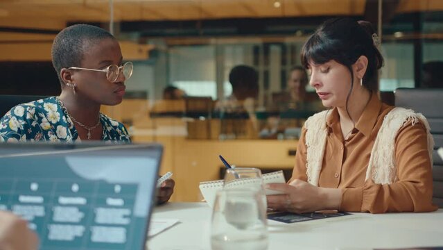 Young Latin businesswoman reading notes in copybook and having conversation with Black female colleague at meeting table in coworking room