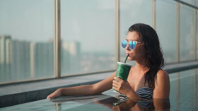 Gorgeous Asian fit Woman drinking cocktail in swimming pool. Water drops flow down tanned body. Girl wearing sunglasses relaxing on vacation at luxury hotel resort. Beautiful slim lady in bikini