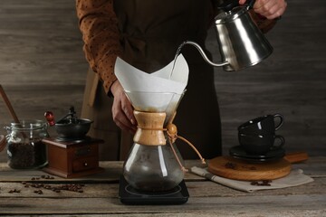 Woman pouring hot water into glass chemex coffeemaker with paper filter at wooden table, closeup