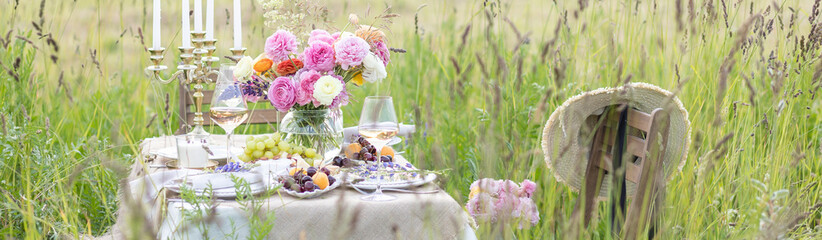 Elegant wedding or private party for a loving couple table setting outdoors in the garden. Beautiful decor with fresh ranunculi flowers, candles. Gourmet cheese board, fruits, white wine. Banner