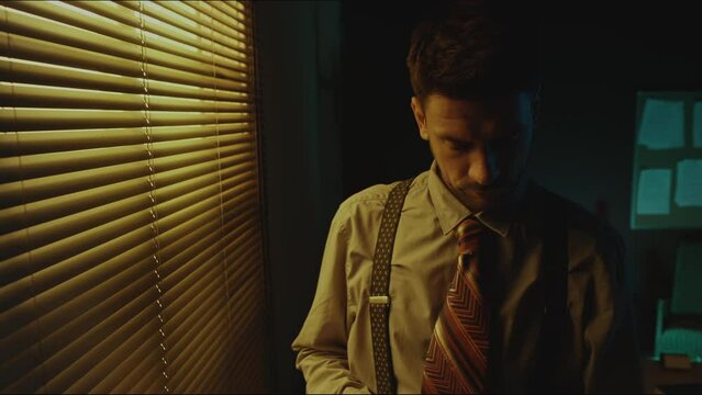 Tilt shot of bearded Caucasian man in vintage office wear looking at pictures standing near window in dark room and then pushing blinds looking out for police and feeling nervous