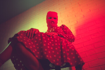 Portrait of a woman in a balaclava sitting on a chair against a brick wall in red neon light.