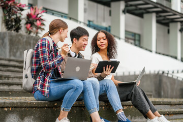 Happy diverse university students sitting on steps, using laptops and tablets, enjoying conversation. Student exchange and study abroad program, Asian man, african american woman and caucasian people.