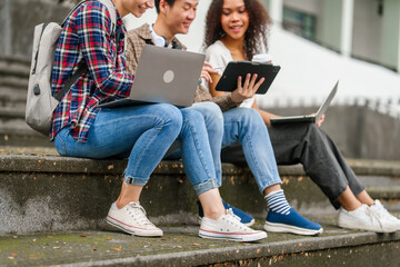 Happy diverse university students sitting on steps, using laptops and tablets, enjoying conversation. Student exchange and study abroad program, Asian man, african american woman and caucasian people.