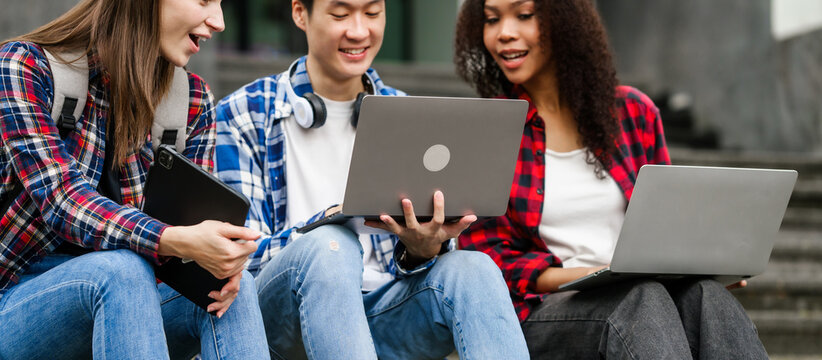 Happy diverse university students sitting on steps, using laptops and tablets, enjoying conversation. Student exchange and study abroad program, Asian man, african american woman and caucasian people.