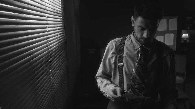 Monochrome medium shot of Caucasian cautious bearded man in retro clothes looking at photographs and pushing blinds peeking outside and standing pensively in room