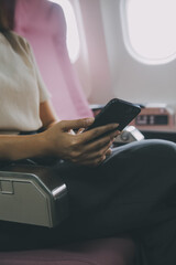 Asian woman passenger sitting in airplane near window and reading news from social networks or using travel applications in smartphone
