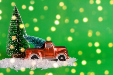 A red pickup truck is carrying a Christmas tree with artificial snow on a green background with bokeh. Banner, New Year card with copy space.