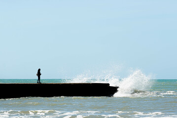 Persona parada contemplando el mar justo en el momento donde una ola grande choca con las rocas. - obrazy, fototapety, plakaty
