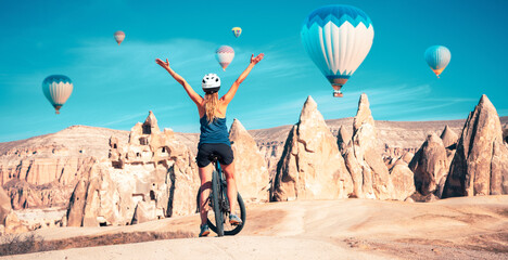 happy woman on bike enjoying view of colorful hot air balloons flying over fairy chimneys valley in Goreme, Cappadocia, Turkey