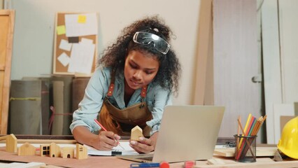 Carpenter america black woman curly hair sketch work and making notes in work paper at desk with laptop computer in wood carpentry workshop, young female learning project online at woodshop
