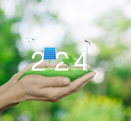 2024 white text with solar cell, wind turbine and growing tree on green grass field in hands over...