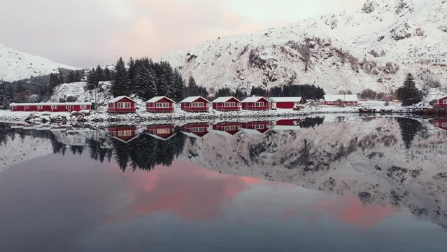 Peaceful winter view of a traditional fishing village, Lofoten Islands, Norway, adorned with red rorbu houses against snowy surroundings. The tranquil landscape is beautifully reflected in the water.